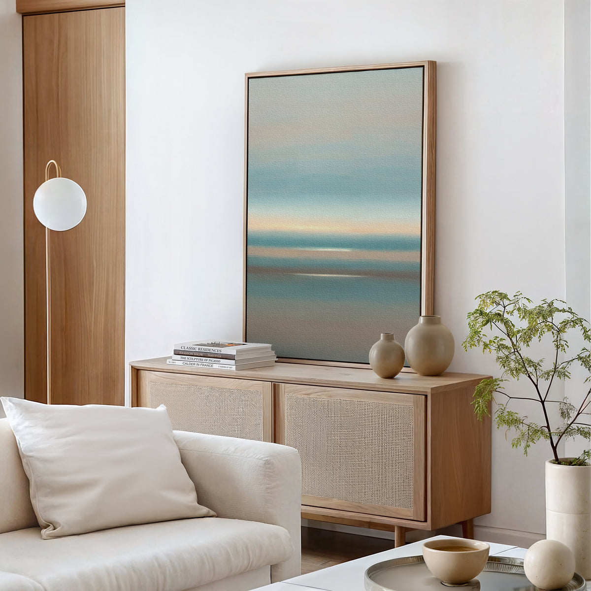 A bright, modern living room features the Soft Tide Layers Canvas Art - Vertical above a light wood sideboard with stacked books, ceramic vases, and a potted plant. A white sofa with a cushion completes the tranquil, minimalist decor.