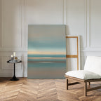 A minimalist room featuring chevron wood floors, a white cushioned chair, a black round table with candles and books, and the Soft Tide Layers Canvas Art - Vertical leaning against a white paneled wall for tranquil home decor.