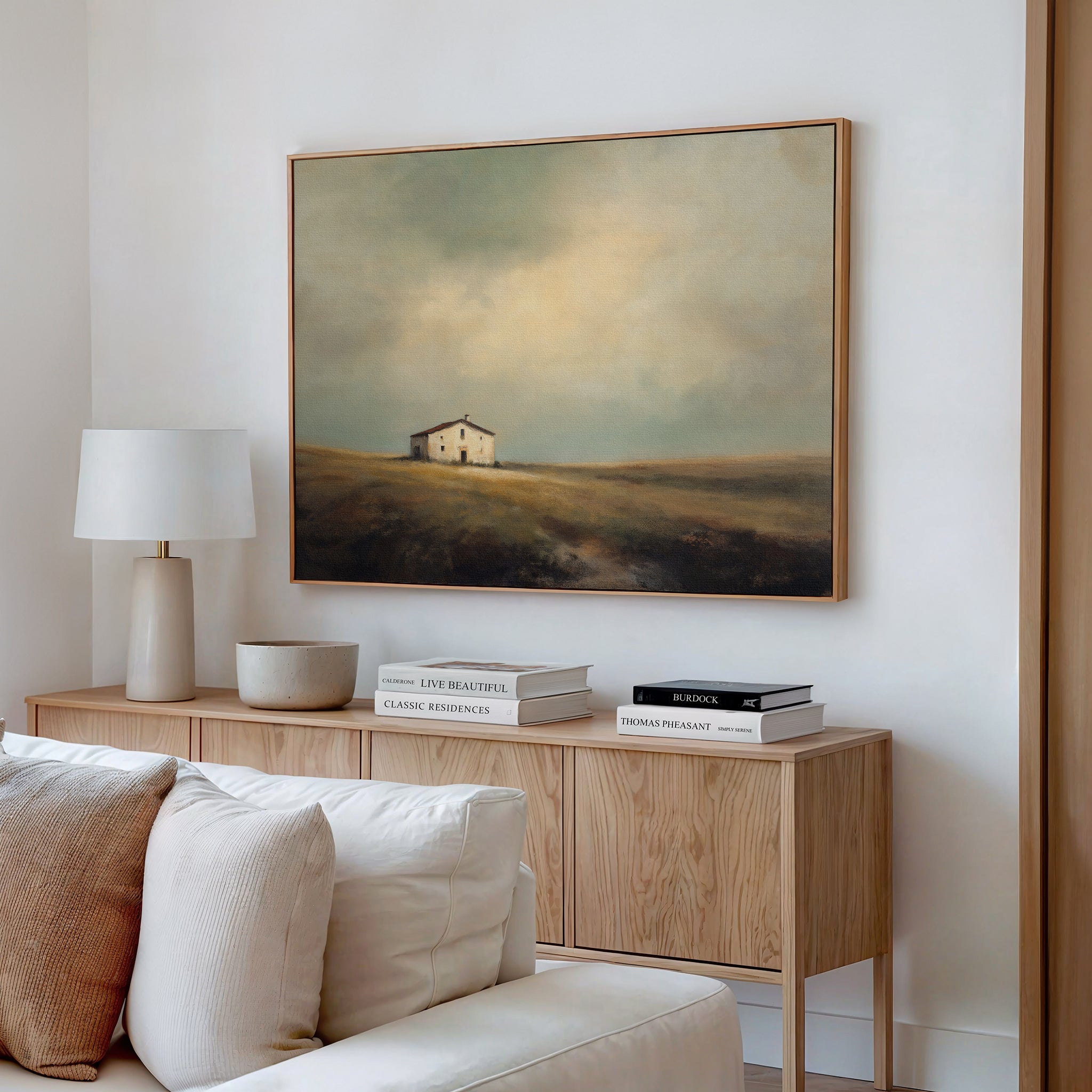 A bright living room features a wood sideboard with stacked books, a lamp, and a beige bowl. Above it hangs the Solitary House Canvas Art - Horizontal, adding rustic charm to the white wall.