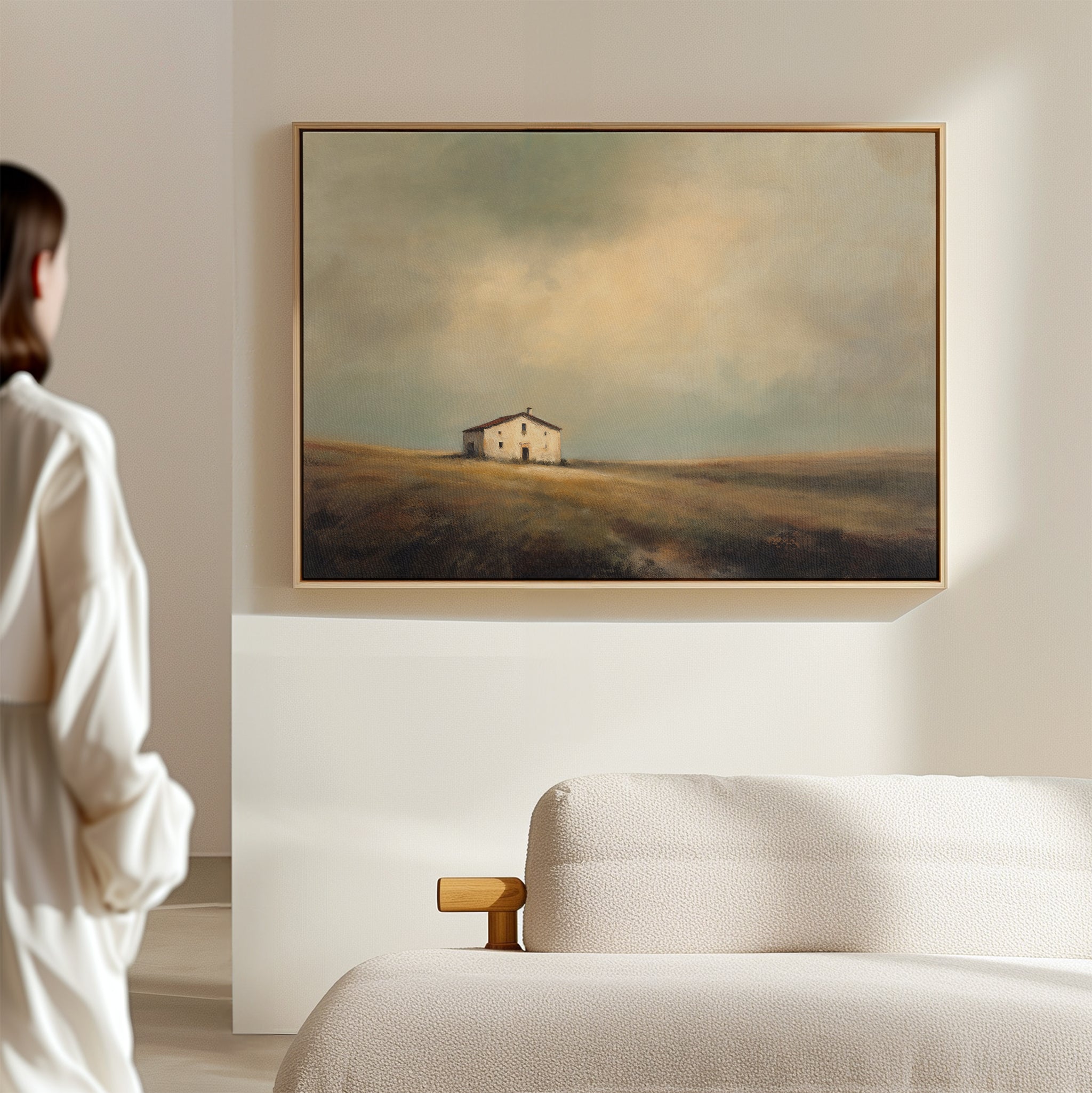 A person stands in a bright, minimal room, admiring the Solitary House Canvas Art - Horizontal depicting a weathered white farmhouse on grassy fields under cloudy skies. A textured beige sofa nearby enhances the rustic charm of the scene.