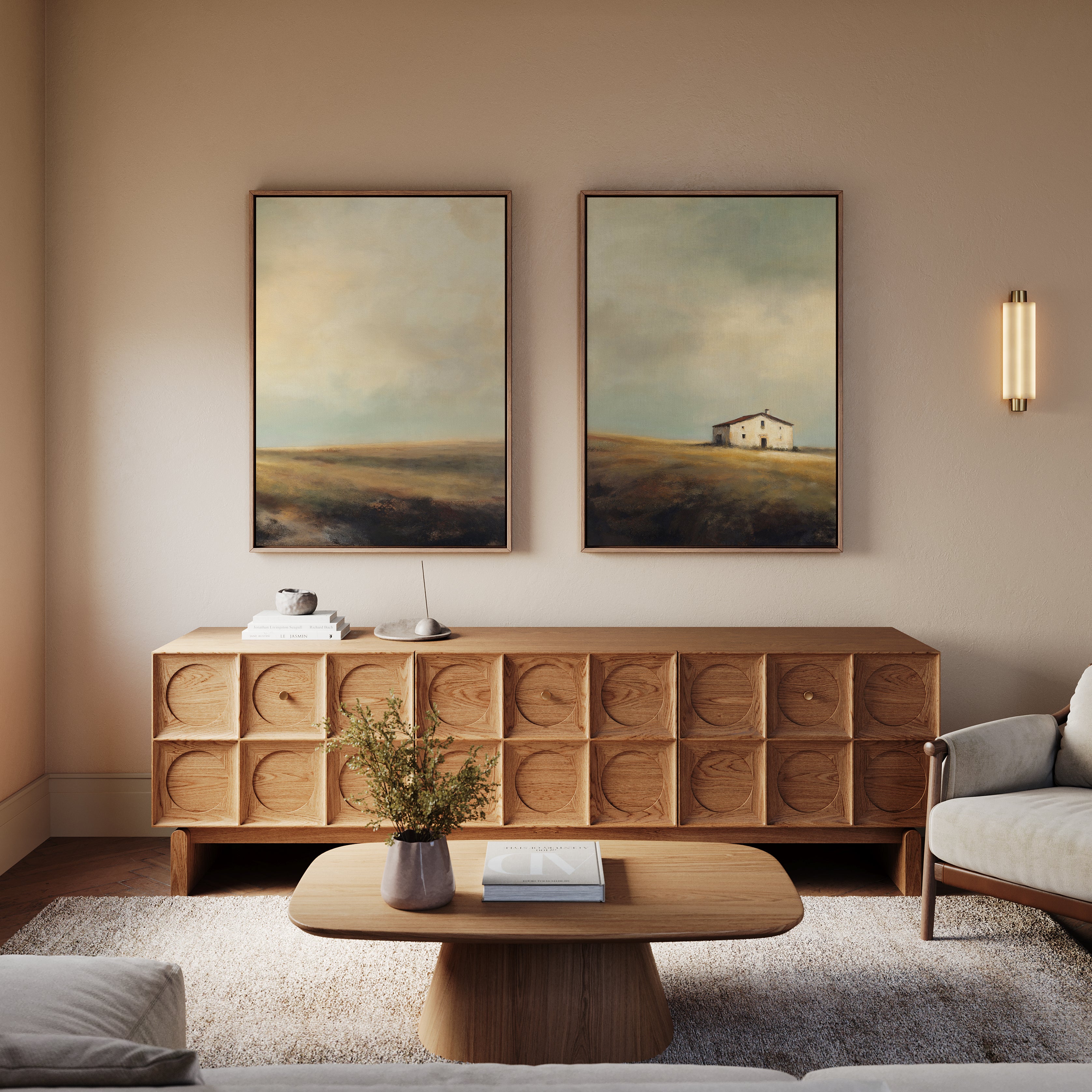 A modern living room with rustic charm features a wooden coffee table, matching sideboard, the Solitary House - Set of 2 landscape paintings, a small sculpture, and a vase—all in soft neutrals for a cozy countryside feel.
