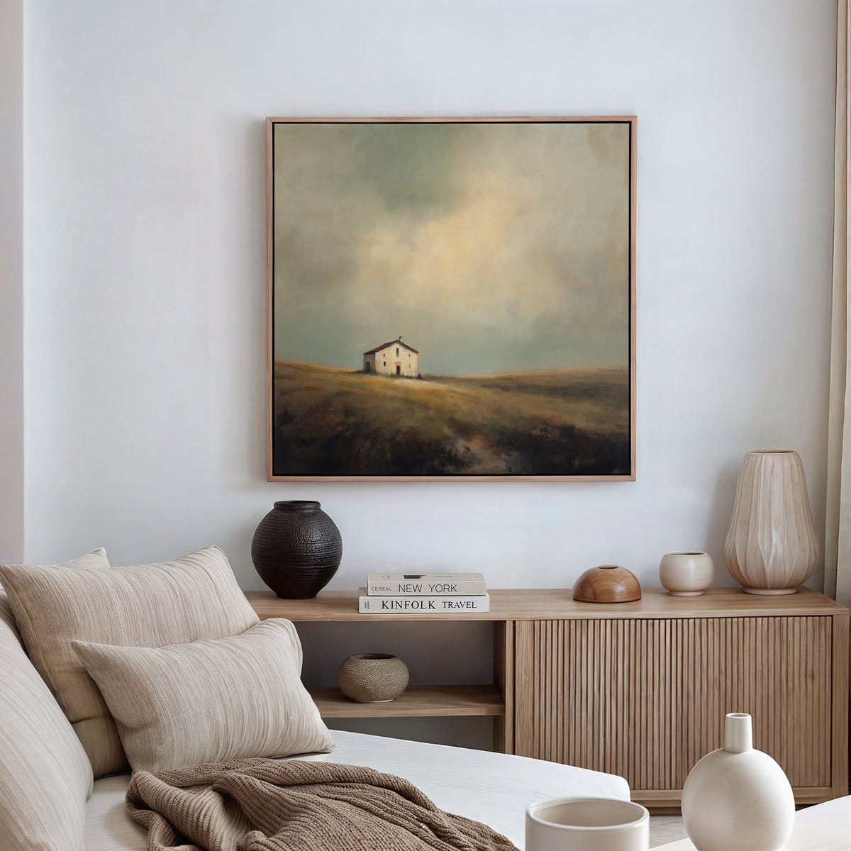A cozy, neutral-toned living room features a beige sofa, wooden accents, and a sideboard topped with books and vases. Above it hangs the Solitary House Canvas Art - Square, depicting a charming rustic house in a misty landscape under a dramatic sky.