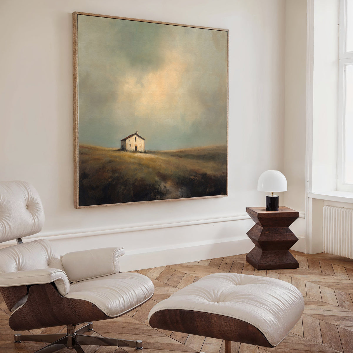 A modern, minimal living room featuring a white lounge chair, ottoman, dark wood side table with a white lamp, and the Solitary House Canvas Art - Square depicting a weathered farmhouse in mist, adding rustic charm to the space.