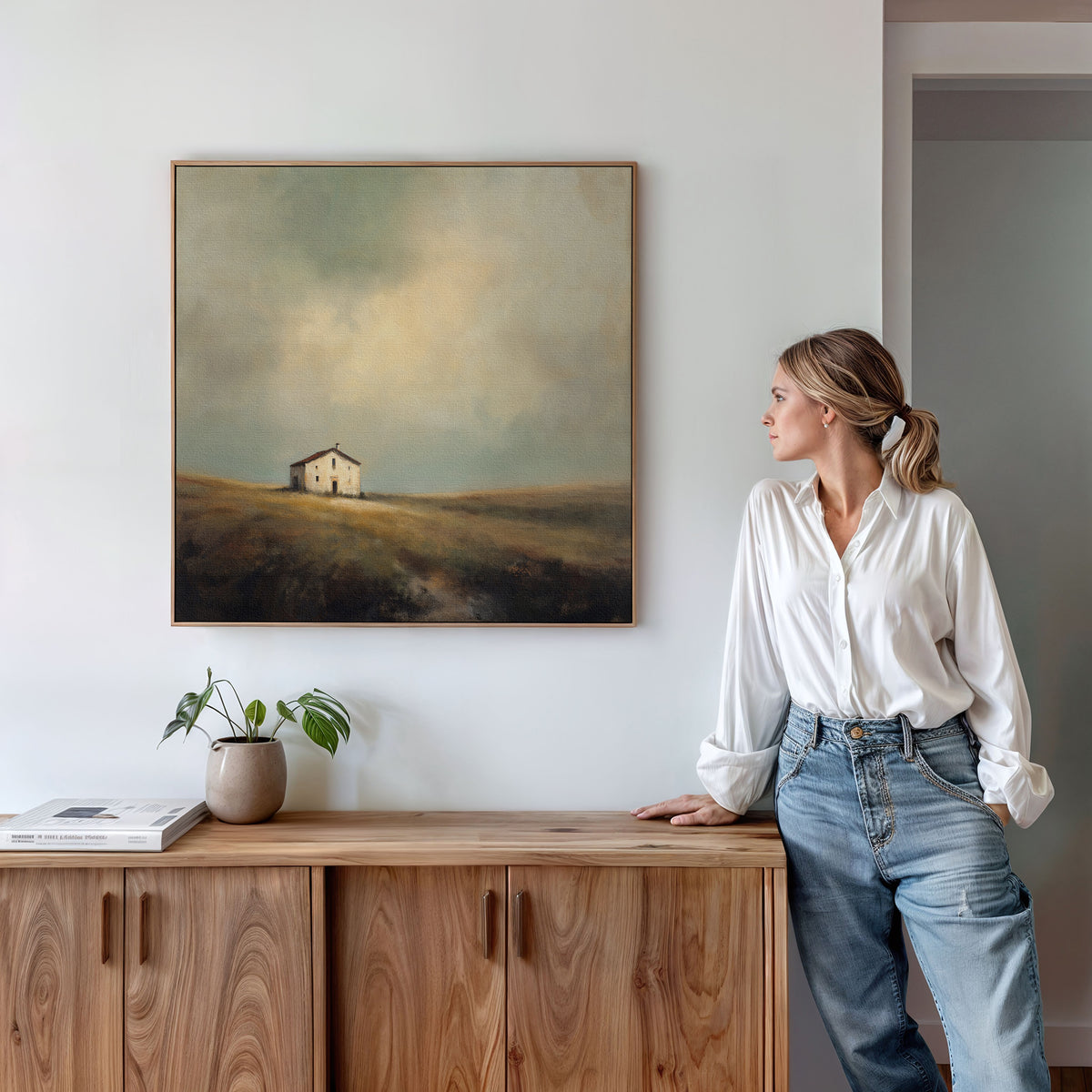 A woman in a white blouse and jeans leans on a wooden cabinet, admiring the Solitary House Canvas Art - Square, which depicts a weathered farmhouse on a grassy hill. Plants and books atop the cabinet enhance the rustic charm of the space.