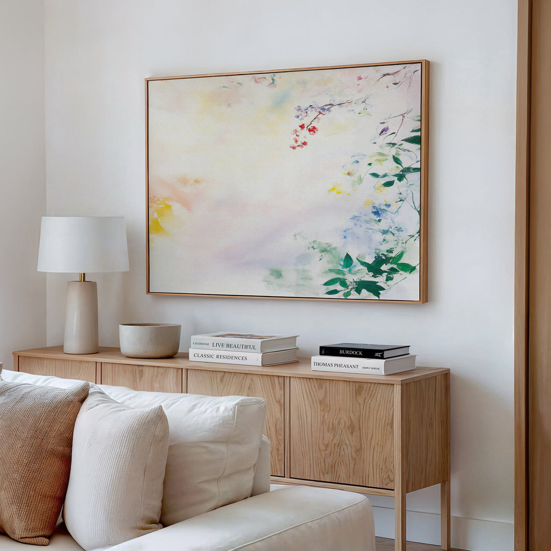 A light wood sideboard with books and a lamp sits against a white wall, topped by Spring Reverie Canvas Art - Horizontal in pastel florals. A white couch is partially visible in the foreground, adding to the airy scene.