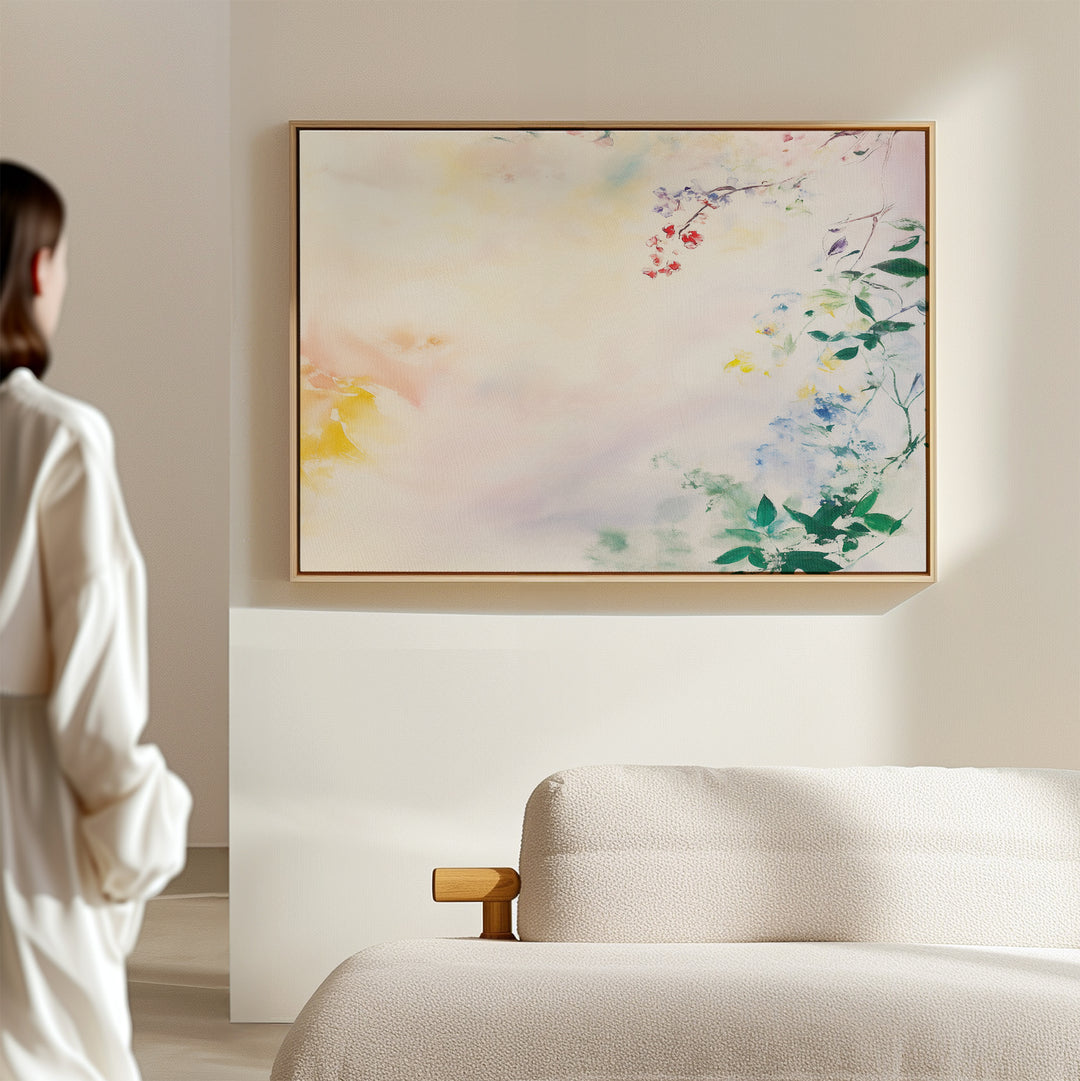 A person in a white shirt admires the Spring Reverie Canvas Art - Horizontal, featuring pastel florals displayed above a cream-colored sofa in a softly lit, minimalist room.