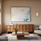 A cozy living room with a light sofa, wooden coffee table, textured sideboard, and the Spring Reverie Canvas Art - Horizontal featuring abstract florals above. A wall sconce and a vase with flowers complete the inviting decor.
