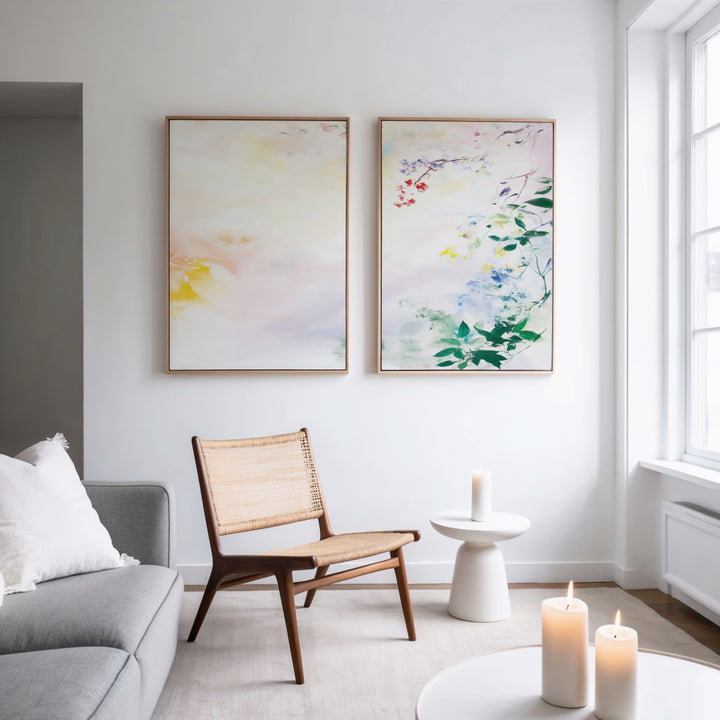 A modern, minimal living room with a wooden chair and small round table holding three lit candles. Above the chair, the "Spring Reverie - Set of 2" abstract watercolor art hangs on a white wall beside a sunlit window.
