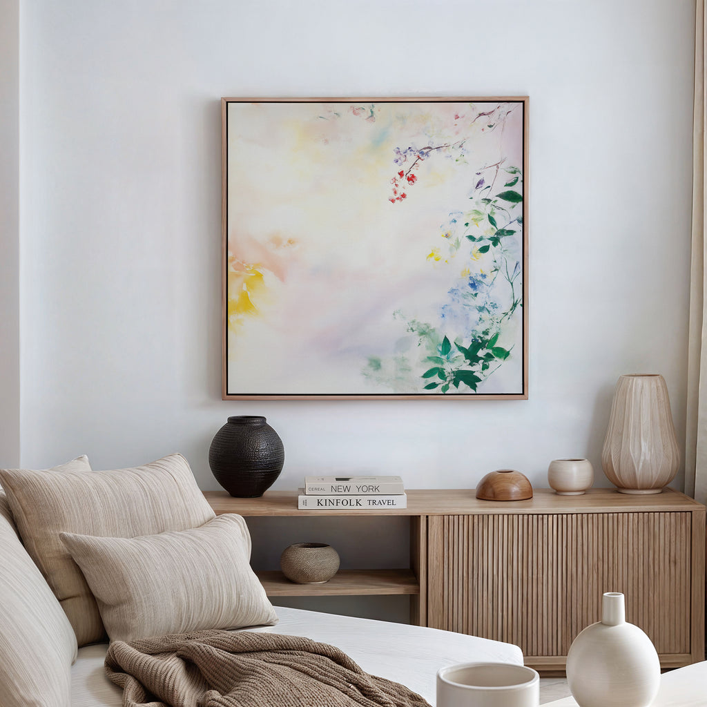 A cozy, neutral-toned living room with a beige sofa, cream cushions, wooden sideboard, vases, books, and the Spring Reverie Canvas Art - Square—a pastel floral piece—adding the perfect touch of decor to the white wall.