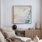 A cozy, neutral-toned living room with a beige sofa, cream cushions, wooden sideboard, vases, books, and the Spring Reverie Canvas Art - Square—a pastel floral piece—adding the perfect touch of decor to the white wall.