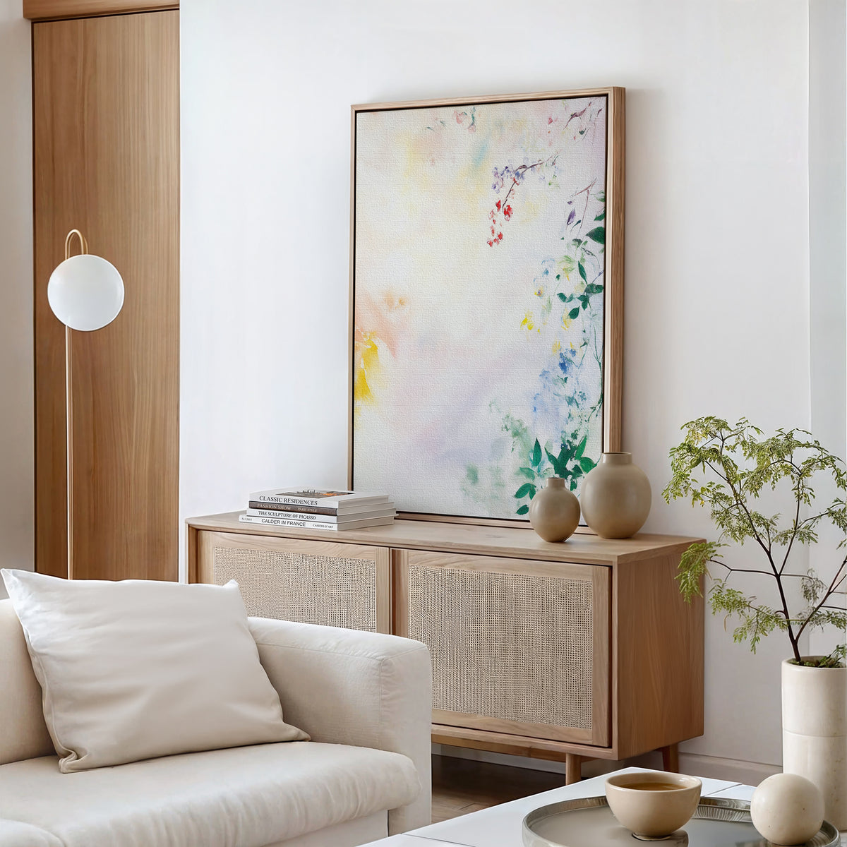 A modern, minimal living room with a cream sofa, light-wood sideboard, Spring Reverie Canvas Art - Vertical above, neutral vases, stacked books, a small green plant, and a round floor lamp set against a white wall.