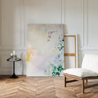 The "Spring Reverie Canvas Art - Vertical," featuring soft pastel florals, leans against a white paneled wall beside an empty frame, with a cushioned chair and round side table on a herringbone wood floor.