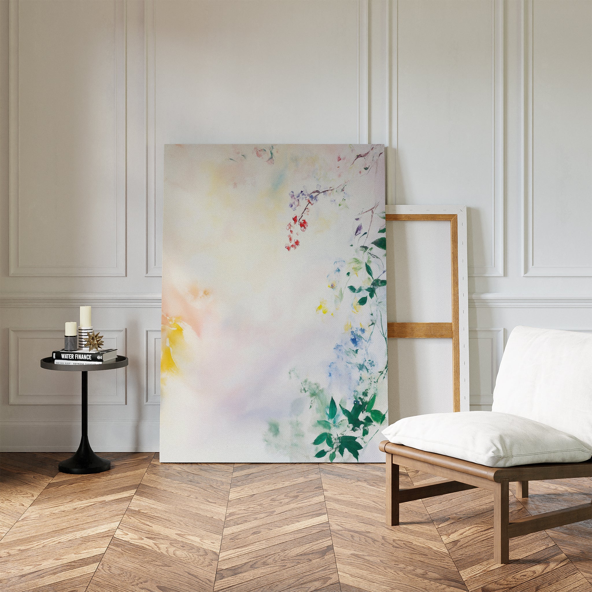 The "Spring Reverie Canvas Art - Vertical," featuring soft pastel florals, leans against a white paneled wall beside an empty frame, with a cushioned chair and round side table on a herringbone wood floor.