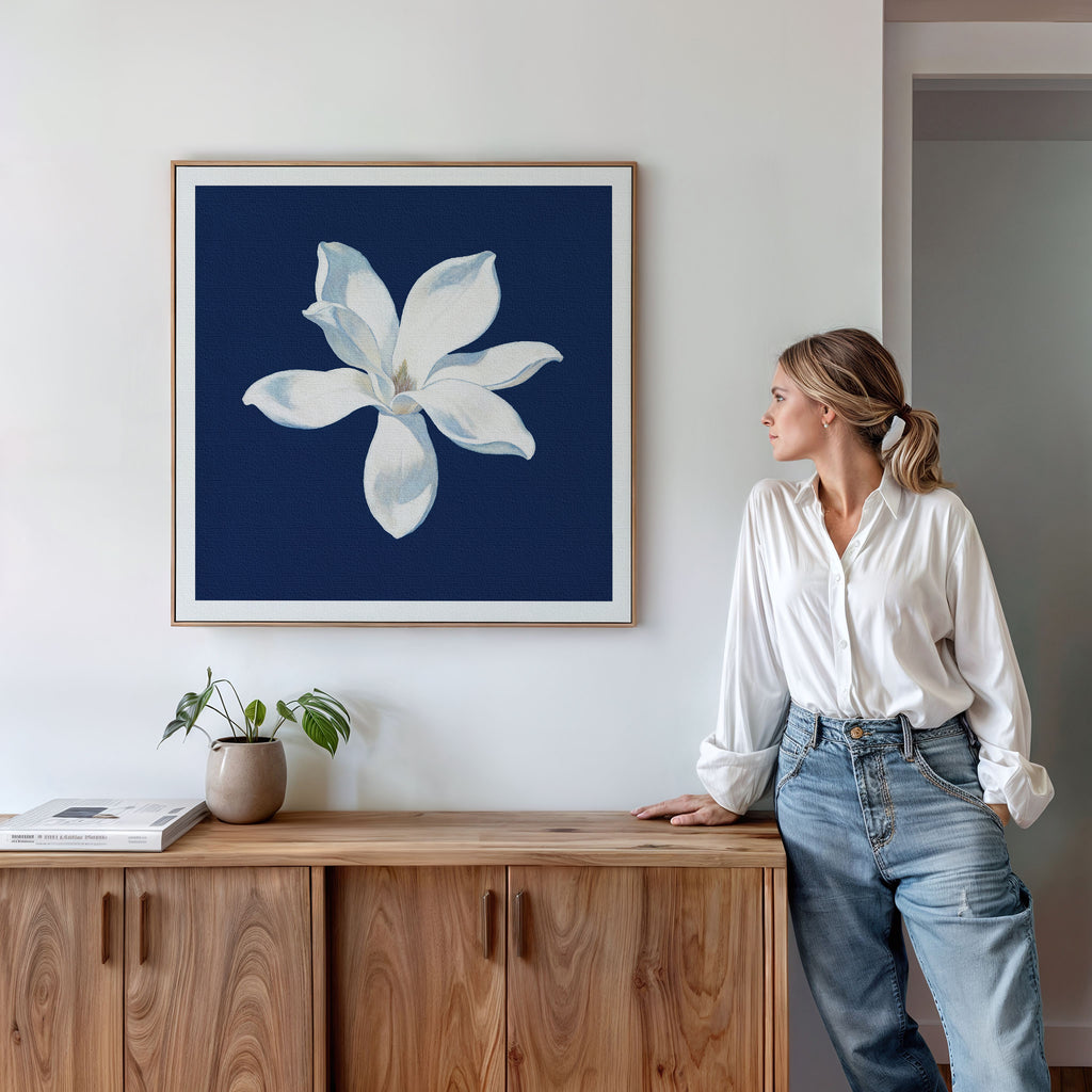A woman in a white blouse and jeans leans against a wooden cabinet, gazing at the Starlit Magnolia Canvas Art - Square, which features a white flower on a dark blue background, hanging above the cabinet.