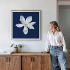 A woman in a white blouse and jeans leans against a wooden cabinet, gazing at the Starlit Magnolia Canvas Art - Square, which features a white flower on a dark blue background, hanging above the cabinet.