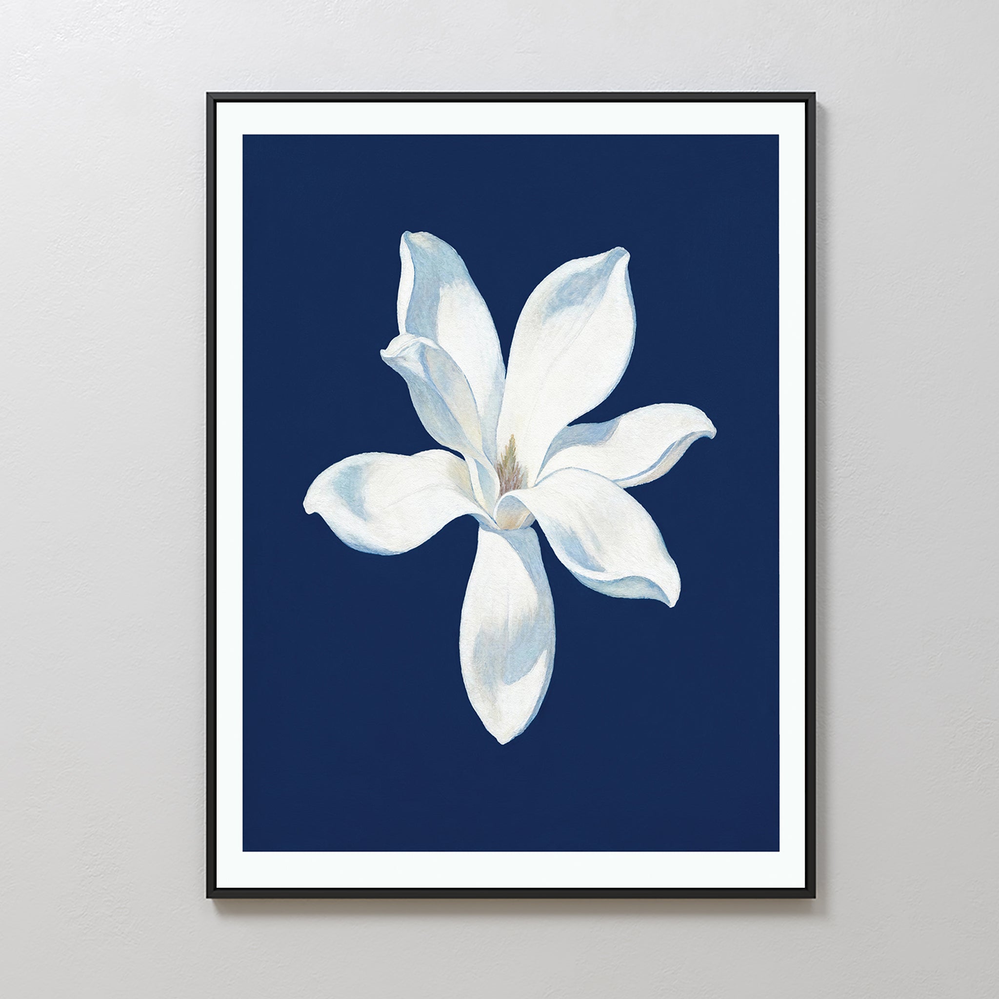 The Starlit Magnolia Canvas Art - Vertical showcases a single white magnolia with soft petals on a deep blue background, framed and displayed on a light gray wall—an ideal piece of contemporary wall art.