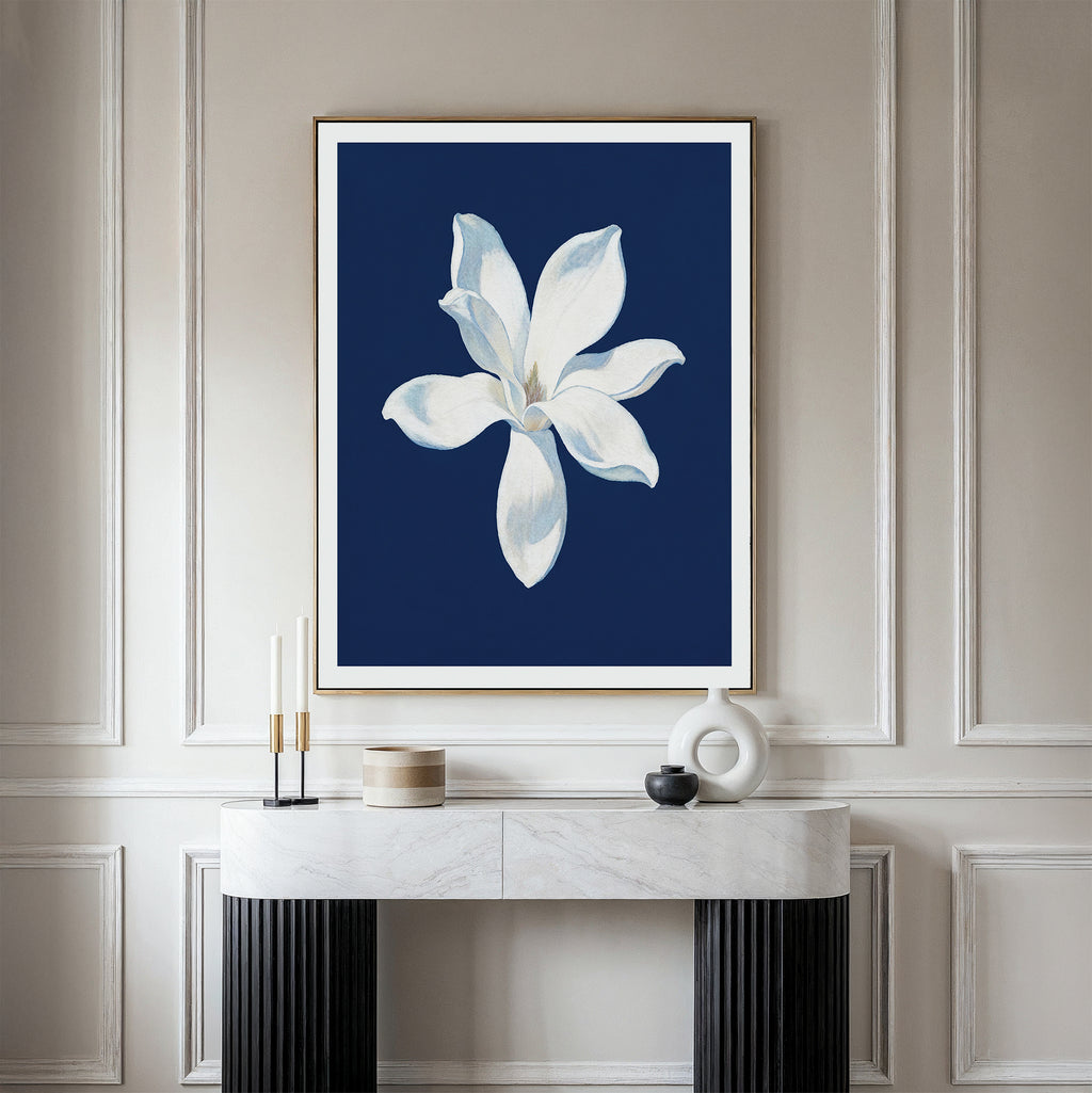 The Starlit Magnolia Canvas Art - Vertical, featuring a white flower on a deep blue background, hangs above a marble console table with modern decor, adding contemporary flair to the light-paneled wall.