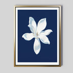 The Starlit Magnolia Canvas Art - Vertical features a single white magnolia flower with subtle shading on a deep blue background, framed in gold—modern wall art ideal for contemporary spaces.
