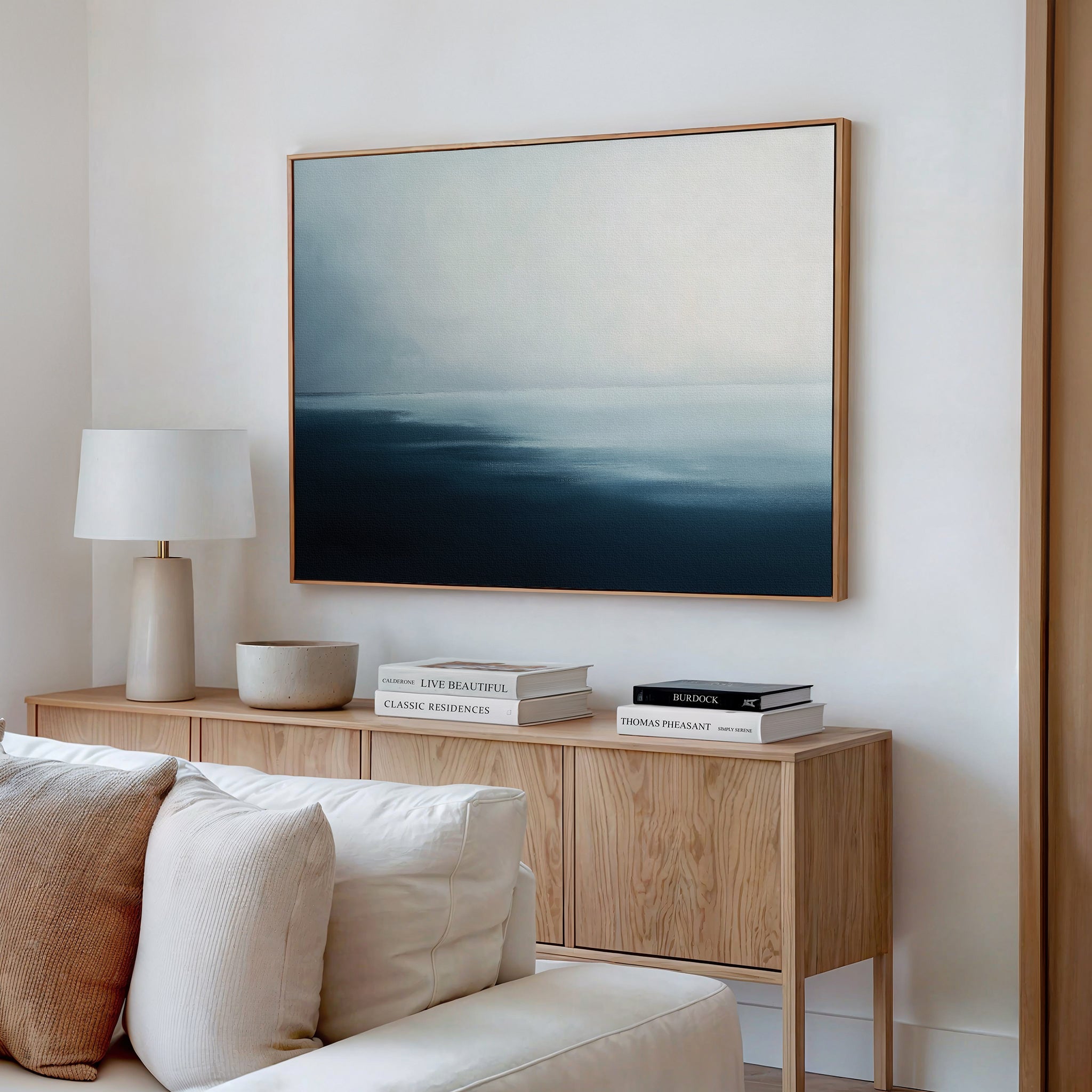 A modern living room featuring a light wood sideboard, stacked books, a white table lamp, a bowl, and the Steel Harbor Mist Canvas Art - Horizontal above a white sofa with neutral cushions creates a serene Nordic Park atmosphere.