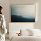 A person in a white shirt stands in a minimalistic room, admiring the "Steel Harbor Mist Canvas Art - Horizontal," a large blue and gray abstract piece displayed above a light-colored sofa.