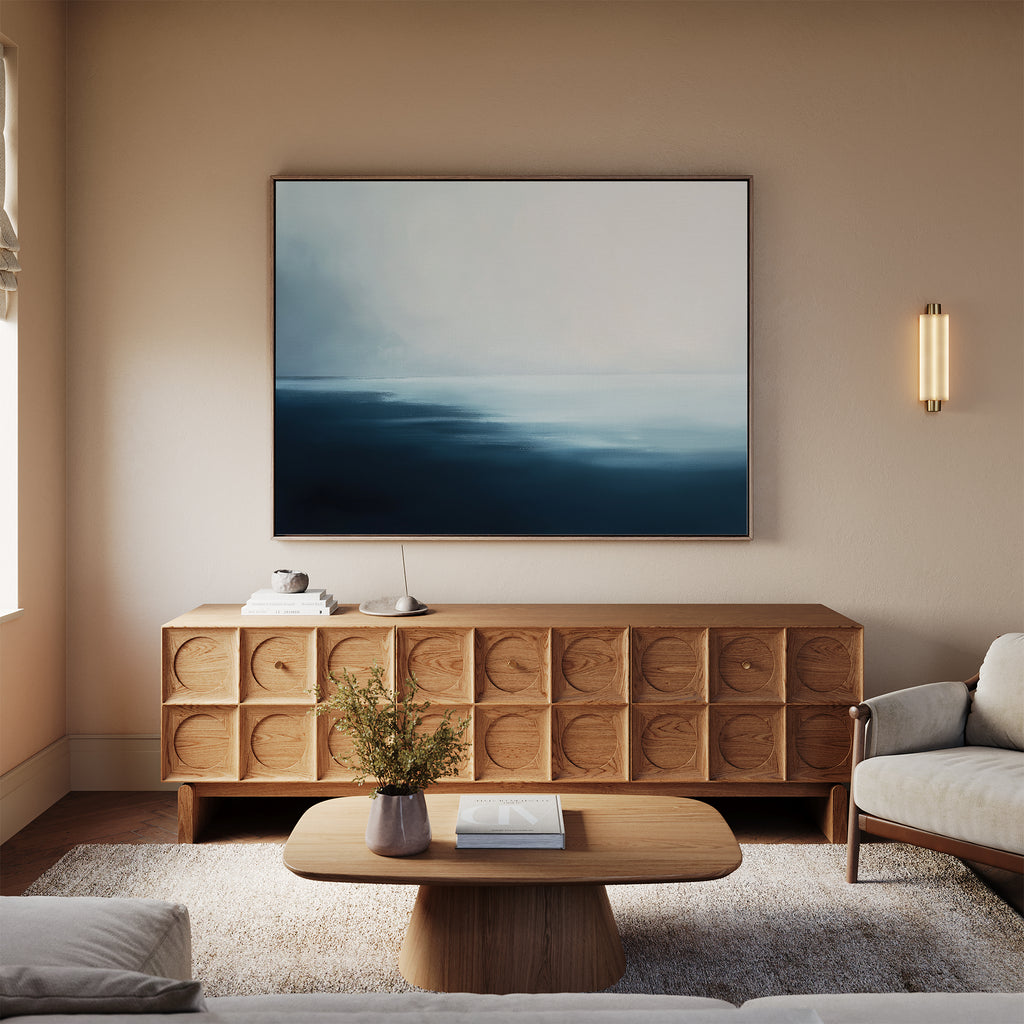 A minimalist living room with a wooden sideboard, Steel Harbor Mist Canvas Art - Horizontal, beige walls, a simple coffee table topped with a vase and two books, a light rug, cream sofa, and gentle wall sconce lighting.