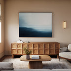 A minimalist living room with a wooden sideboard, Steel Harbor Mist Canvas Art - Horizontal, beige walls, a simple coffee table topped with a vase and two books, a light rug, cream sofa, and gentle wall sconce lighting.