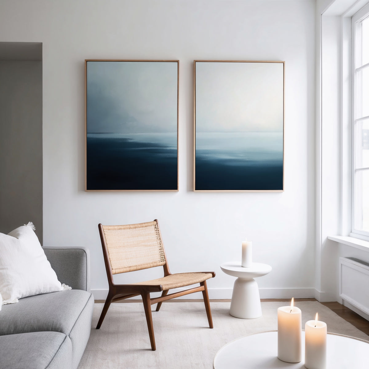 A minimalist living room features a gray sofa, rattan chair, white side table, and three lit candles. Above the chair, the Steel Harbor Mist - Set Of 2 abstract wall art in steel blue hangs on a white wall with natural light from tall windows.