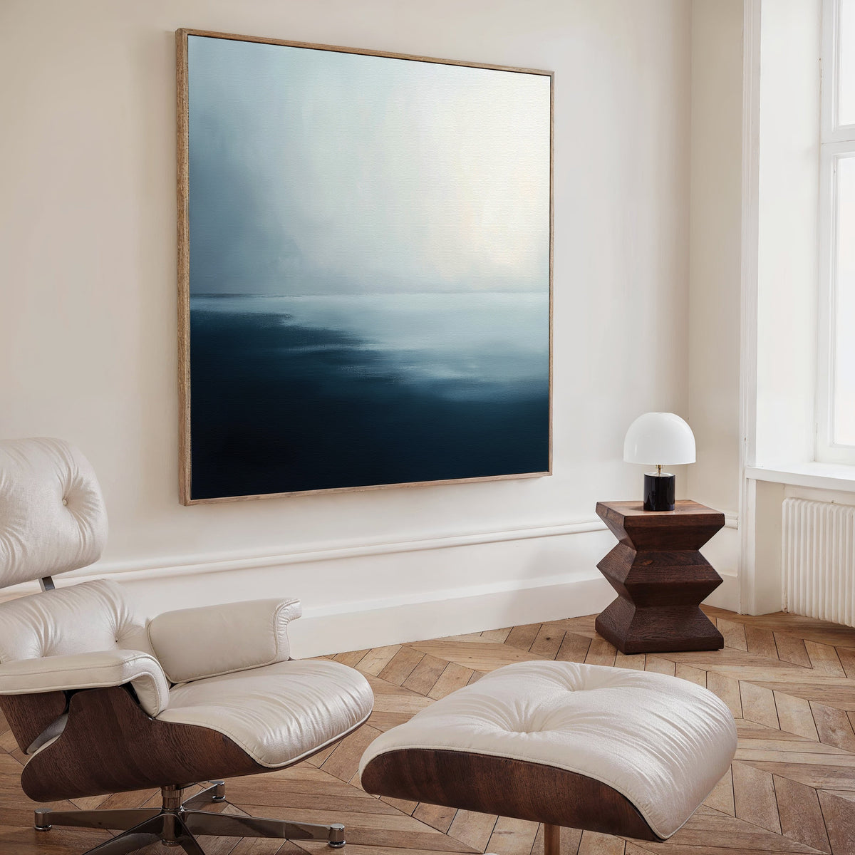 A modern, minimal living room with a white leather lounge chair and ottoman, a zigzag wooden side table with a small lamp, and the Steel Harbor Mist Canvas Art - Square adding striking blue and white tones. Light pours in from tall windows.