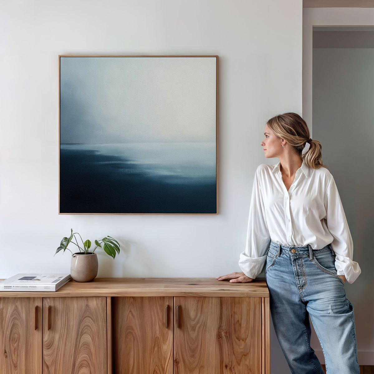 A woman in a white blouse and jeans stands by a wooden sideboard, admiring the Steel Harbor Mist Canvas Art—a minimalist square painting in blue and white. A small plant and book rest on the sideboard, adding to the tranquil scene.