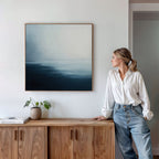 A woman in a white blouse and jeans stands by a wooden sideboard, admiring the Steel Harbor Mist Canvas Art—a minimalist square painting in blue and white. A small plant and book rest on the sideboard, adding to the tranquil scene.