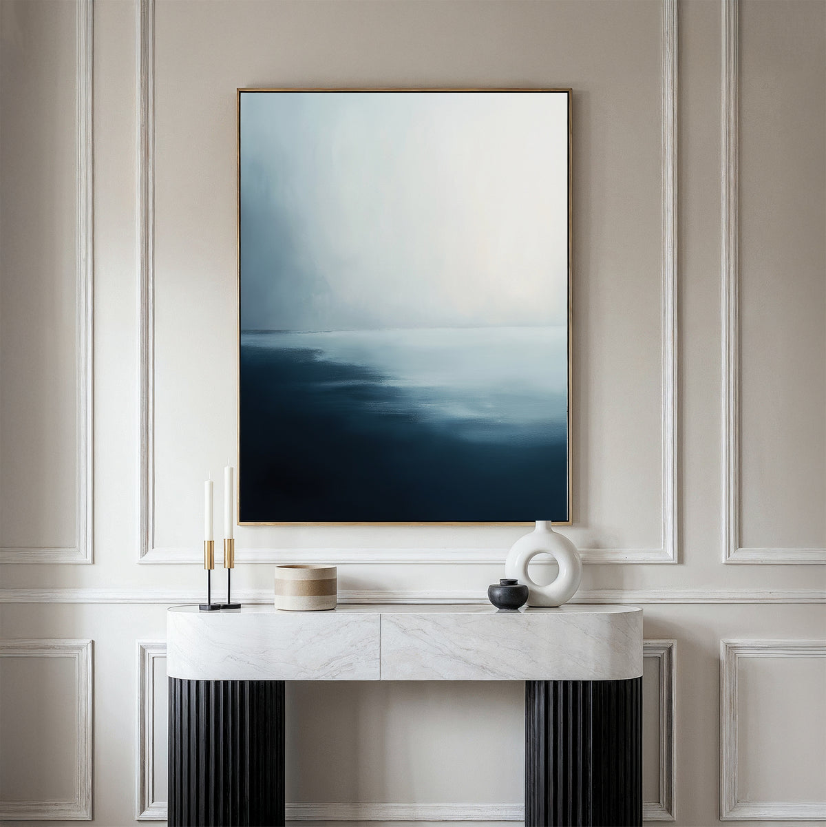 A minimalist living room features a marble console table, decorative objects, and striking wall art—a Steel Harbor Mist Canvas Art - Vertical in blue and white—set against a beige paneled wall.