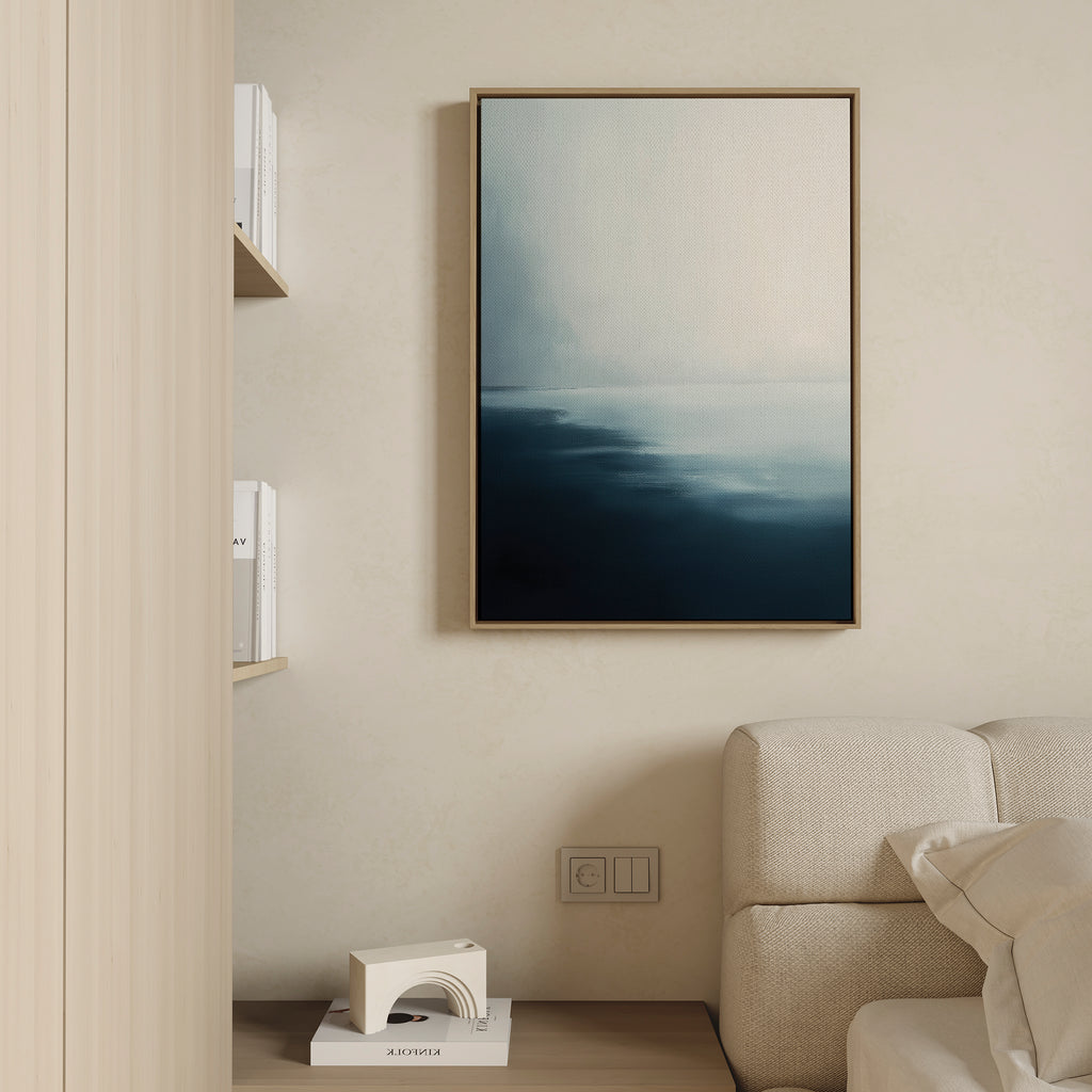 A minimal living room with a light sofa, wooden shelf of white books, and the Steel Harbor Mist Canvas Art - Vertical—an abstract blue and white framed piece. A small geometric decor accent rests on the floor.