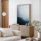 A minimalist living room features the Steel Harbor Mist Canvas Art - Vertical, neutral pottery, stacked books, a light wood sideboard, a white sofa with a cushion, a round lamp, and a small green plant.