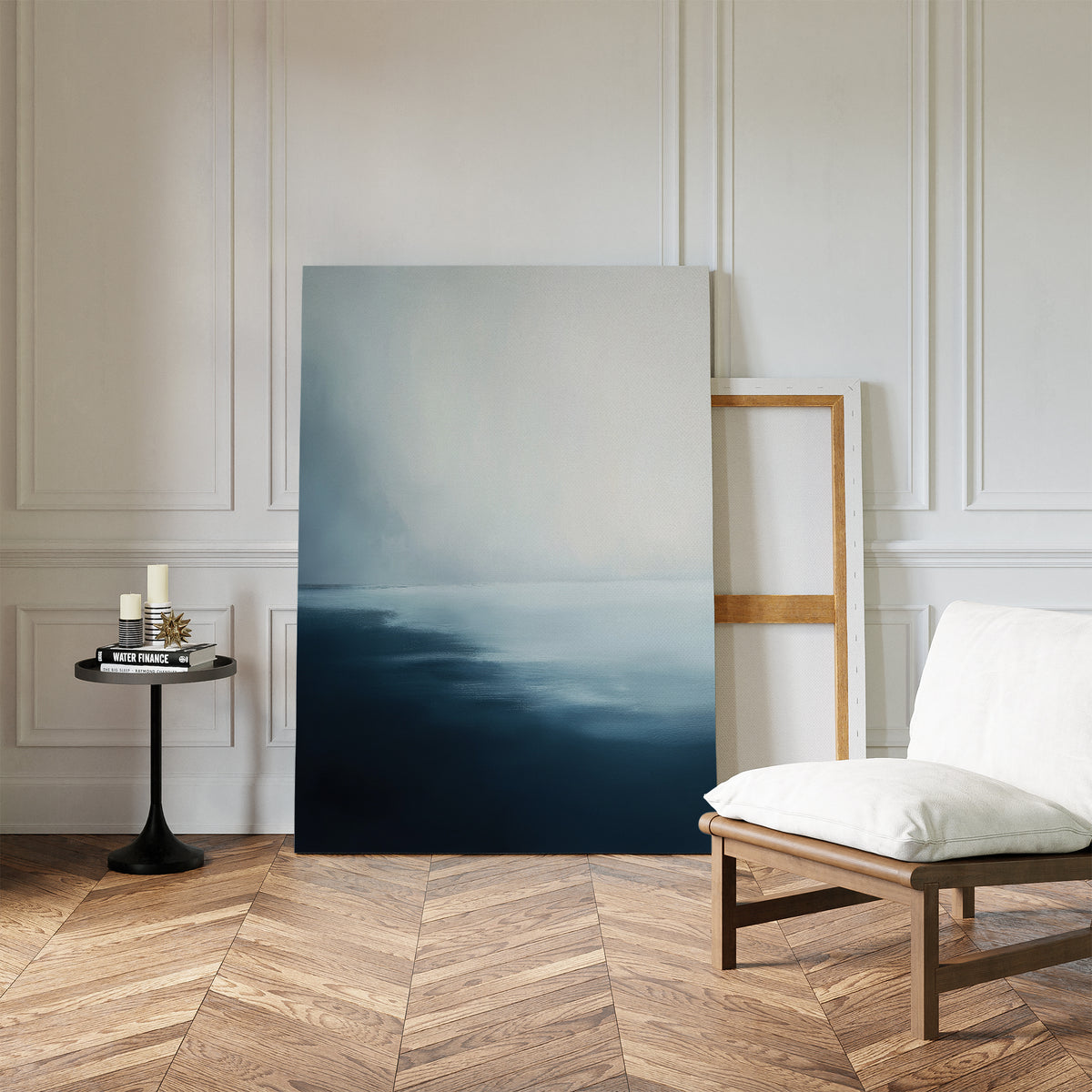 A minimalist living room with herringbone wood floors, a white cushioned chair, round black side table with candles and books, and Steel Harbor Mist Canvas Art - Vertical propped against a white paneled wall.