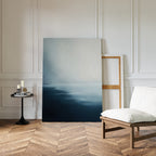 A minimalist living room with herringbone wood floors, a white cushioned chair, round black side table with candles and books, and Steel Harbor Mist Canvas Art - Vertical propped against a white paneled wall.