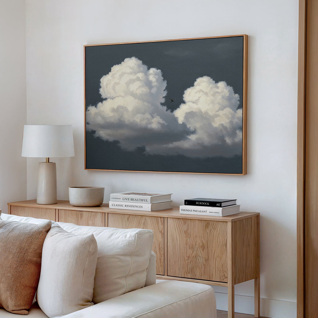 A modern living room with a wooden sideboard, white table lamp, stacked books, beige bowl, and the Storm Passage Canvas Art - Horizontal depicting clouds hanging above on the white wall.