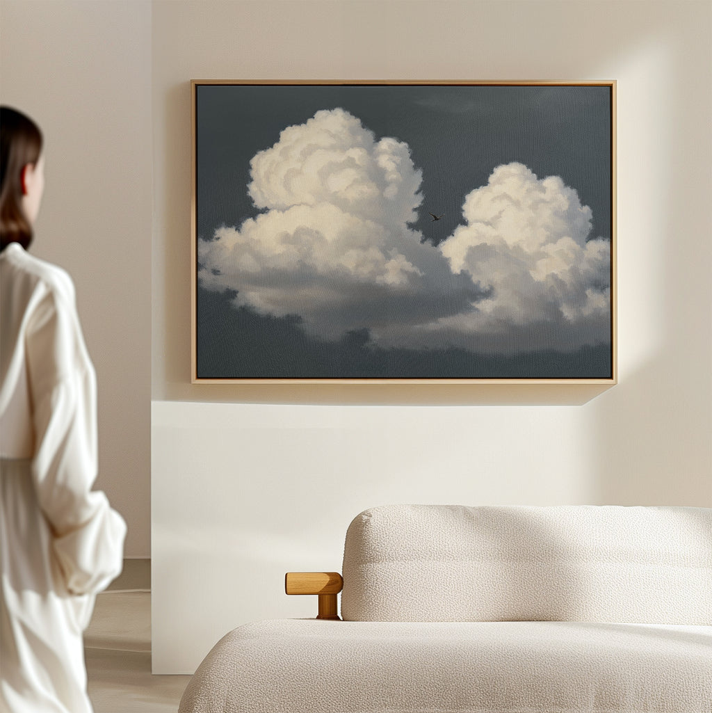 In a bright, minimal room, a woman gazes at the Storm Passage Canvas Art - Horizontal, featuring fluffy clouds and a bird in a dark sky, elegantly displayed above a cream-colored sofa.