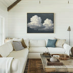 A bright, cozy living room features a white sectional sofa with assorted pillows, a wooden coffee table with decor, and the Storm Passage Canvas Art - Horizontal showcasing fluffy clouds above the couch.