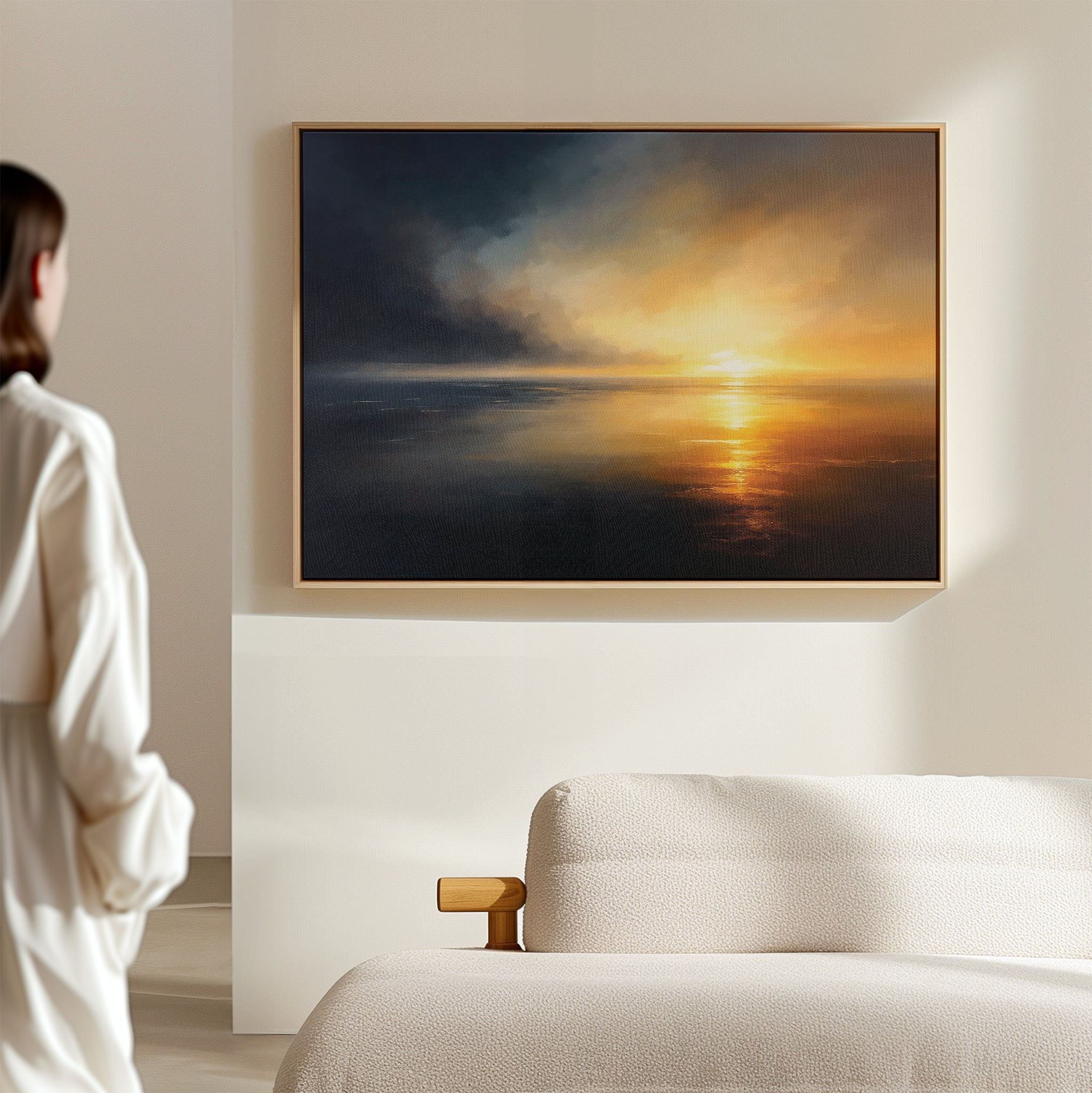 A person in white stands in a modern living room, gazing at the Storm's Edge Canvas Art - Horizontal, featuring a dramatic sunset over water and storm clouds, displayed above the sofa.