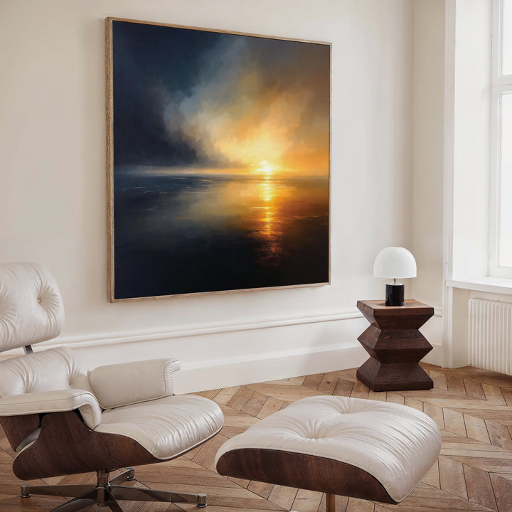 A modern living room with a leather lounge chair and ottoman, a small wooden side table with a white lamp, and Storm's Edge Canvas Art - Square—an atmospheric large canvas print—hanging on the wall.