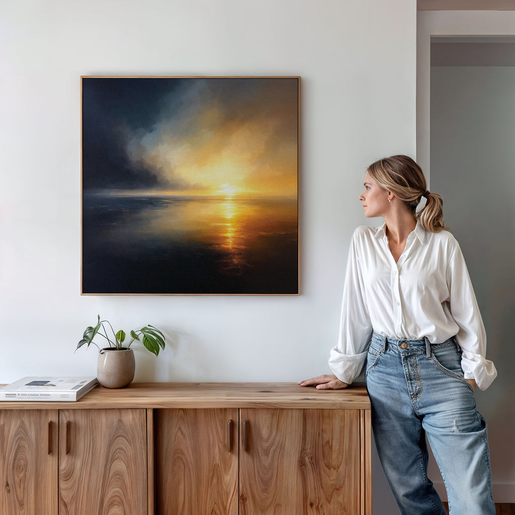 A woman in a white blouse and jeans leans against a wooden cabinet, gazing at the Storm's Edge Canvas Art - Square, which hangs on a white wall. A small plant and a book rest atop the cabinet.