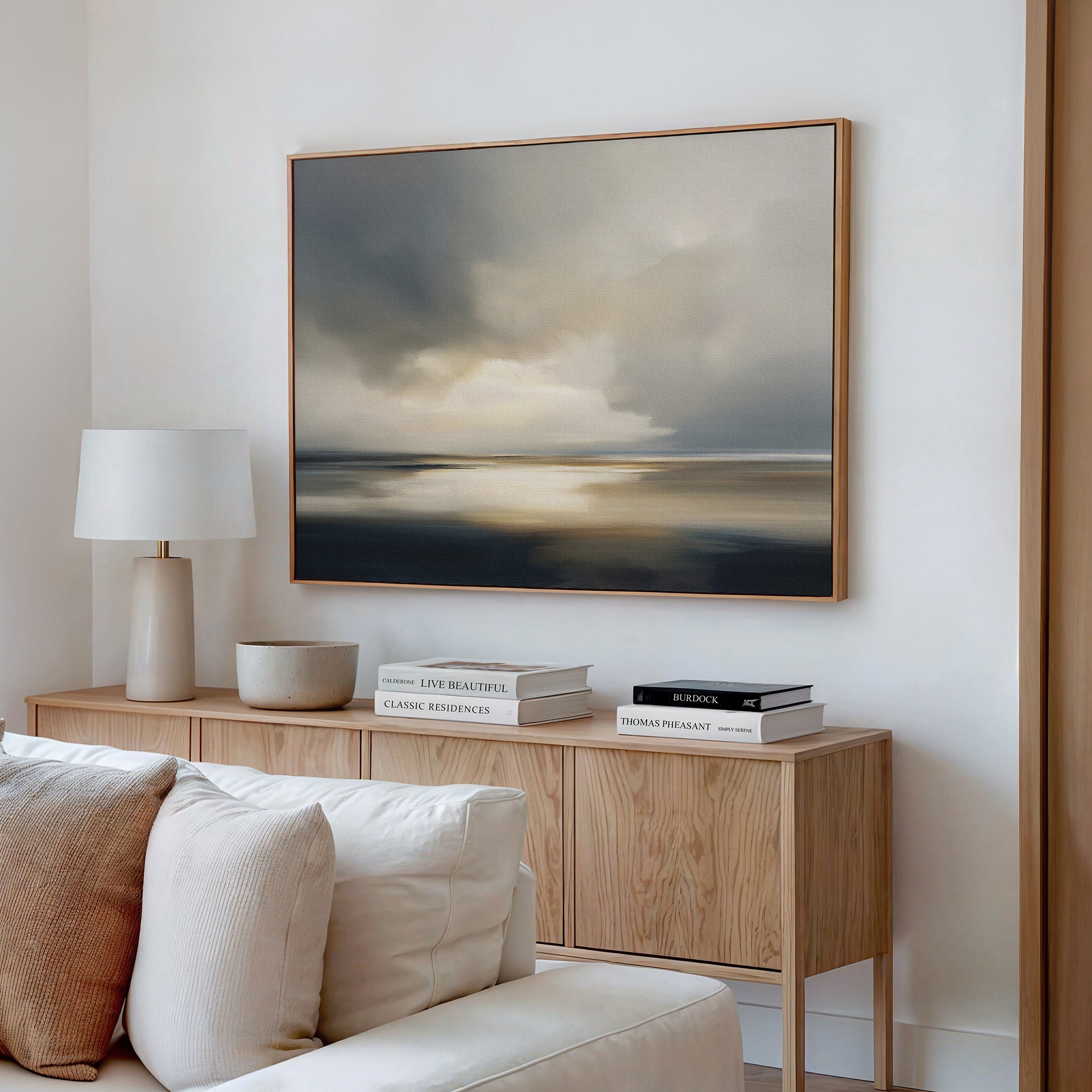 A minimal living room features a light wood sideboard, stacked books, a white lamp, decorative bowls, a cream sofa, and coastal elegance with the Stormy Tides Canvas Art - Horizontal as a large framed abstract landscape painting.