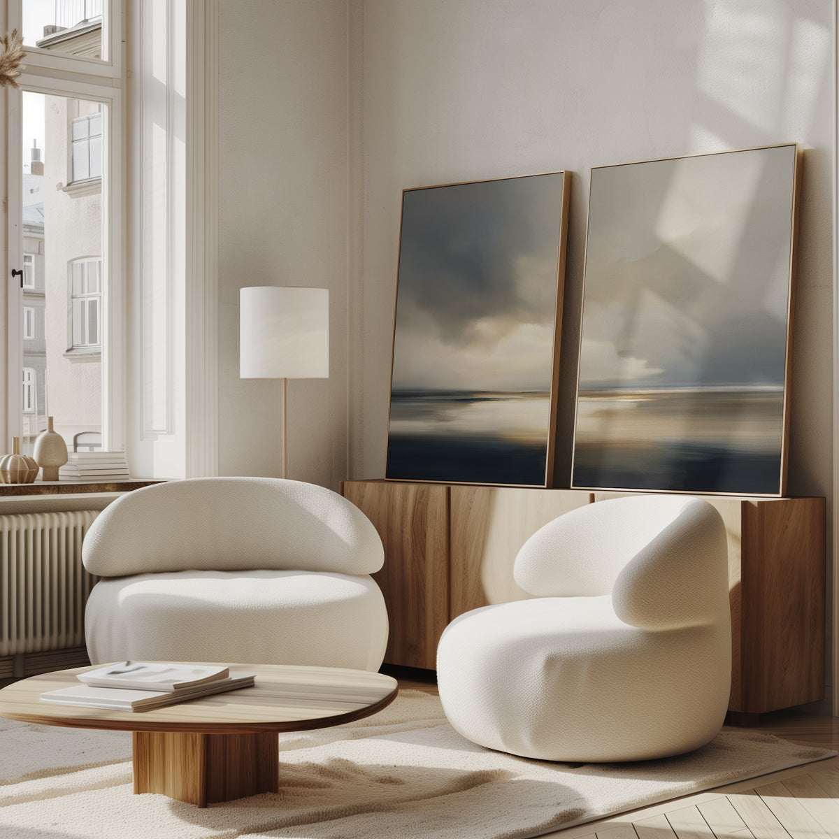 Two modern, rounded white chairs and a low wooden table sit in a sunlit, minimalist living room. Behind them, the "Stormy Tides - Set Of 2" abstract landscape paintings rest on a wooden sideboard against the wall.