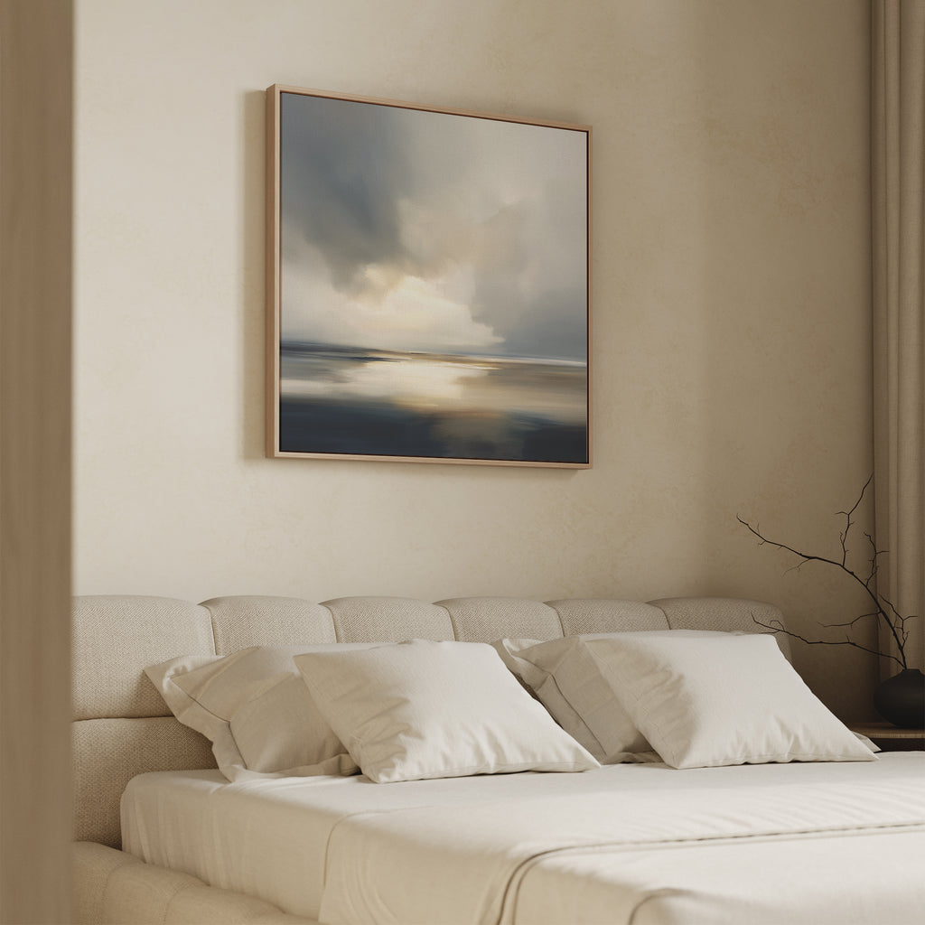 A minimal, airy bedroom features a beige upholstered bed with white bedding and pillows. Above the bed hangs the Stormy Tides Canvas Art - Square, showcasing soft blue, gray, and cream tones. A bare branch decorates the side table.