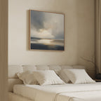 A minimal, airy bedroom features a beige upholstered bed with white bedding and pillows. Above the bed hangs the Stormy Tides Canvas Art - Square, showcasing soft blue, gray, and cream tones. A bare branch decorates the side table.