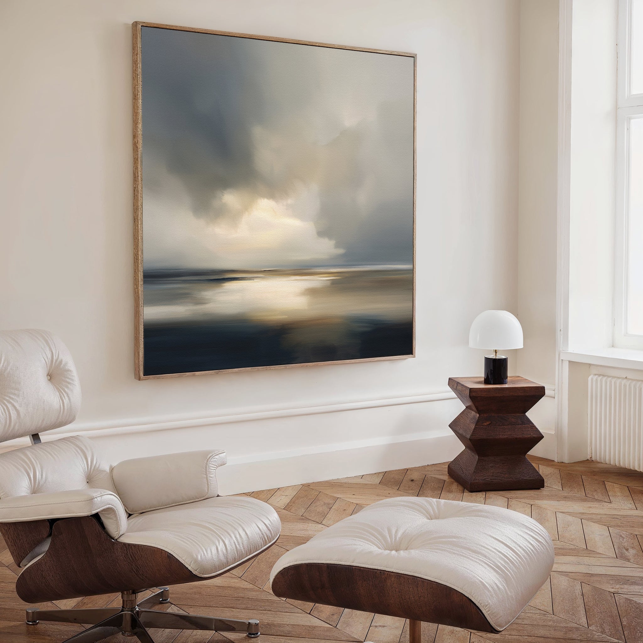 A modern, minimalist room evokes Nordic Park vibes with a cushioned lounge chair and ottoman, geometric wooden side table with a white lamp, and the Stormy Tides Canvas Art - Square featuring moody sky and sea tones on a light wall.