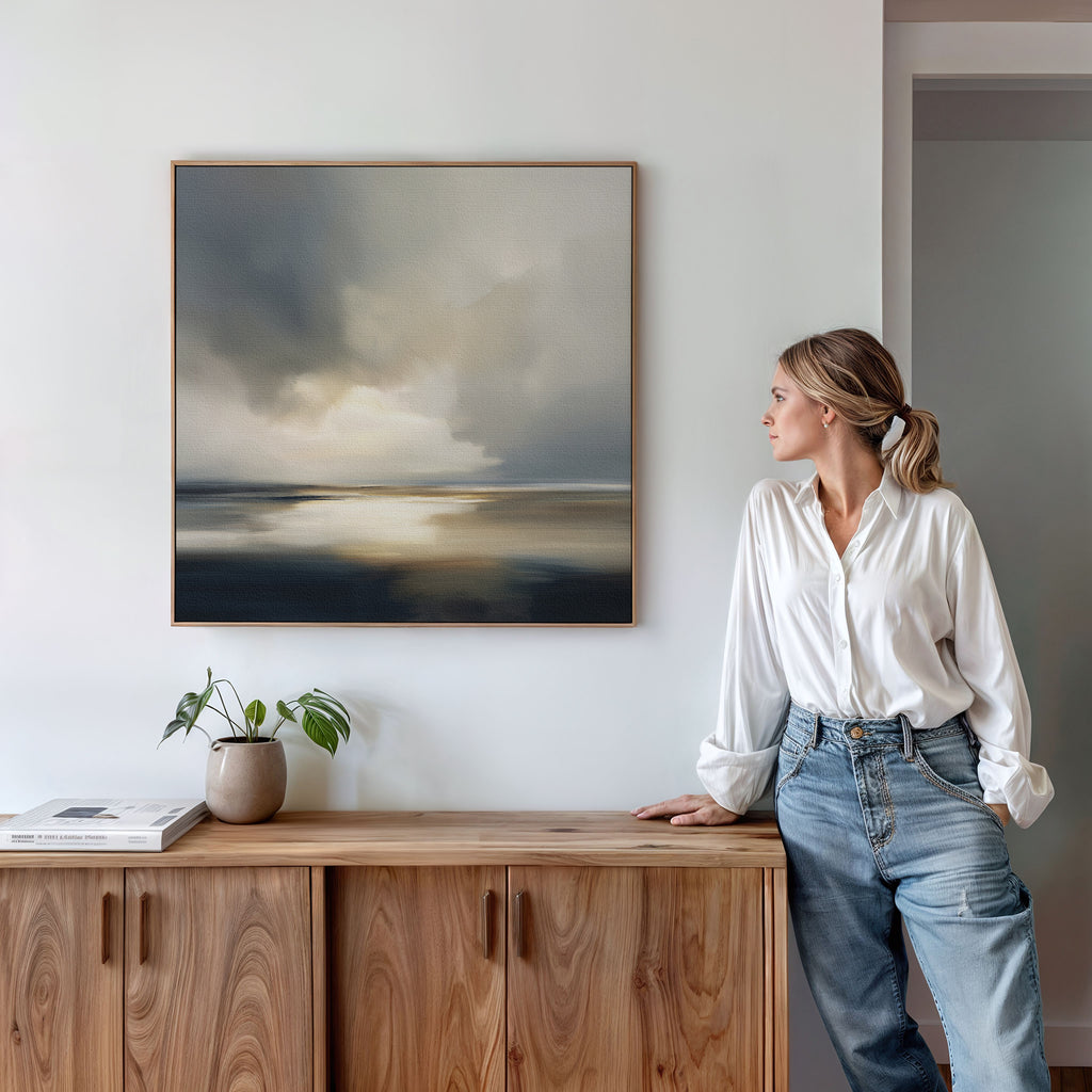 A woman in a white blouse and jeans stands by a wooden sideboard, gazing at the Stormy Tides Canvas Art - Square on a white wall. A small potted plant and books rest on the sideboard, enhancing this Nordic Park-inspired interior.