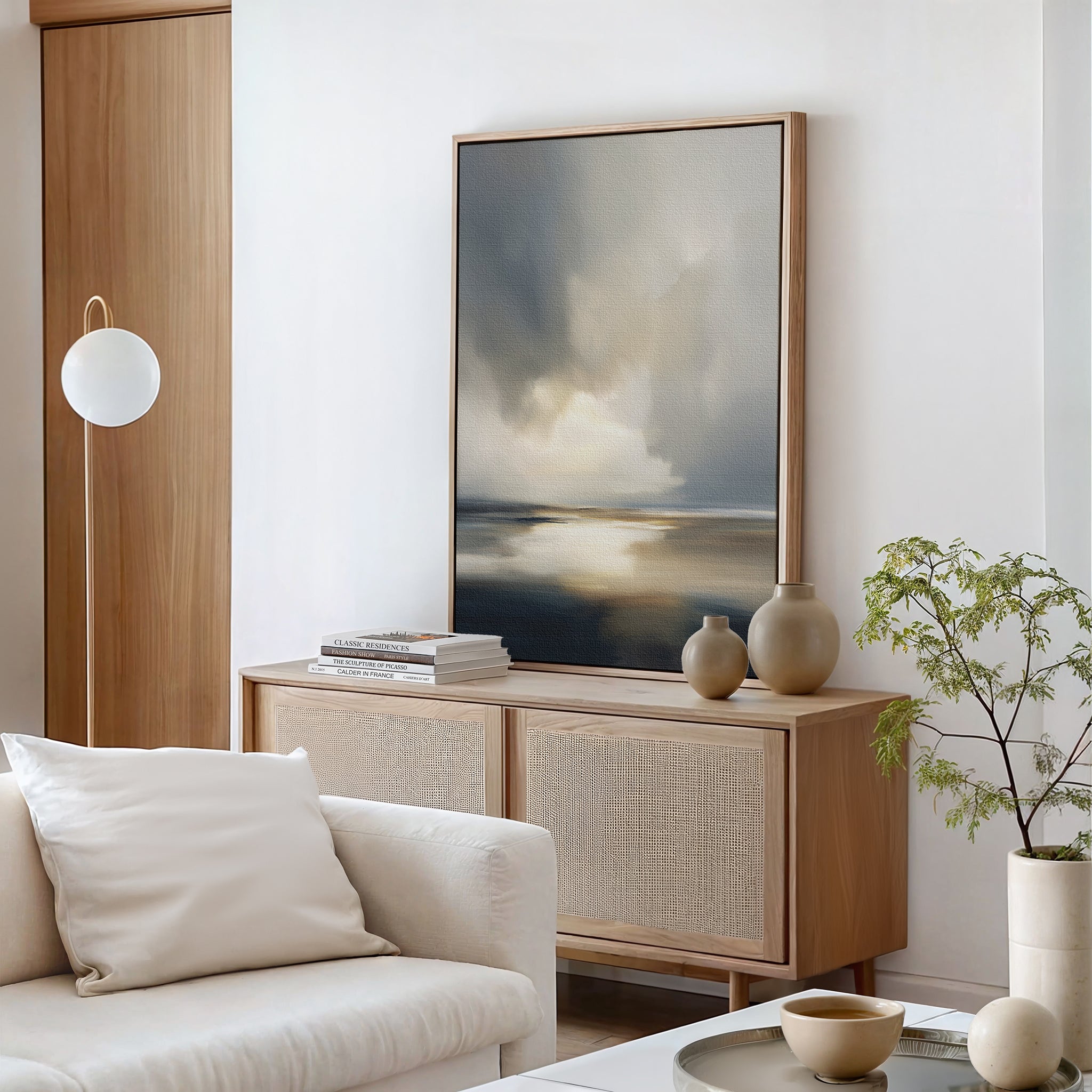A modern, minimalist living room features a light wood sideboard, neutral vases, books, Stormy Tides Canvas Art - Vertical above a white sofa with pillows, a round lamp, and a small potted plant—perfect for a Nordic Park-inspired retreat.