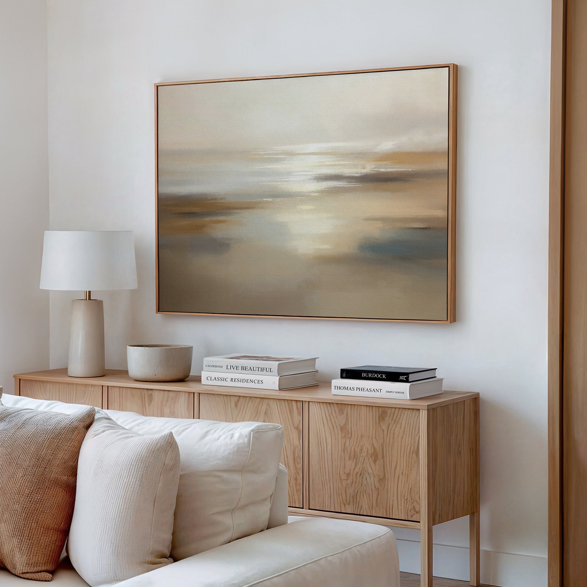 A minimalist living room featuring a light wood sideboard with stacked books, a ceramic lamp, a bowl, and the "Sunlit Waters Canvas Art - Horizontal"—a large abstract seascape in soft beige and gray—on the white wall above.