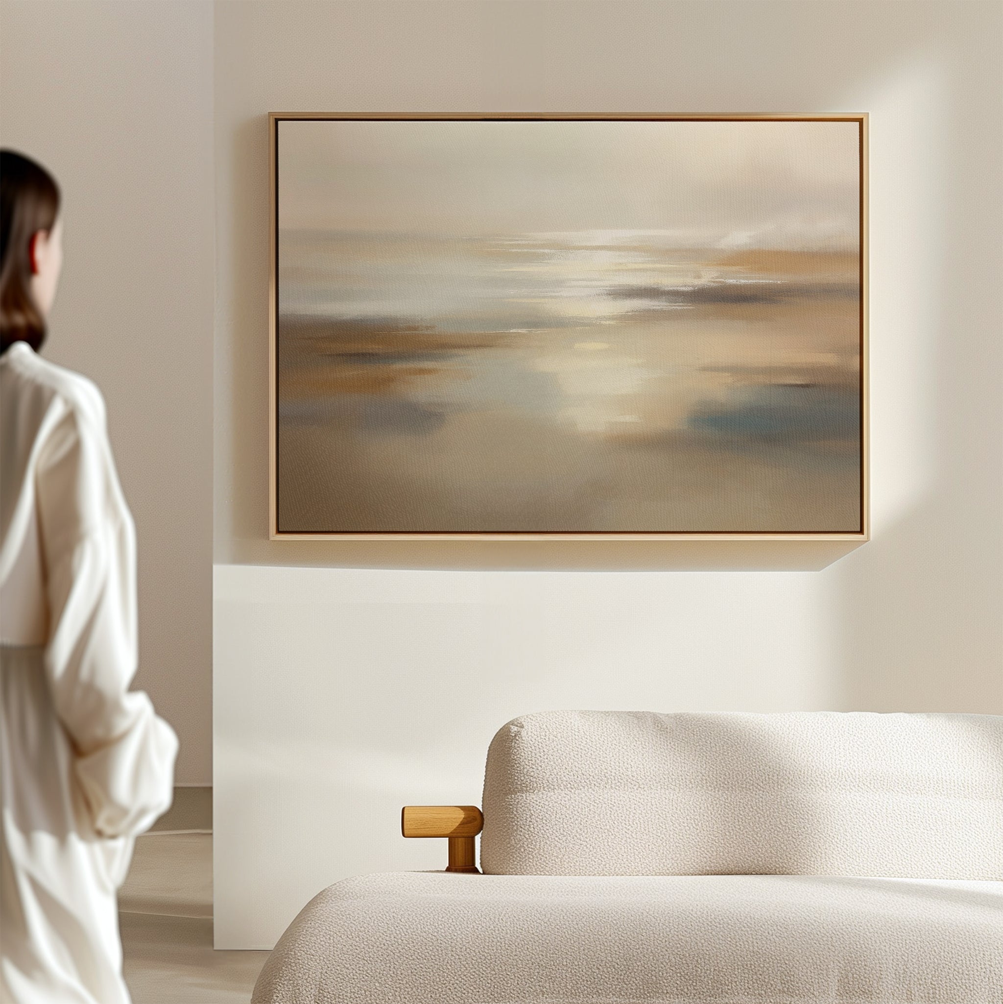 A person in a white robe stands in a minimal, softly lit room, gazing at the Sunlit Waters Canvas Art - Horizontal in beige and blue tones above a textured, light-colored sofa.
